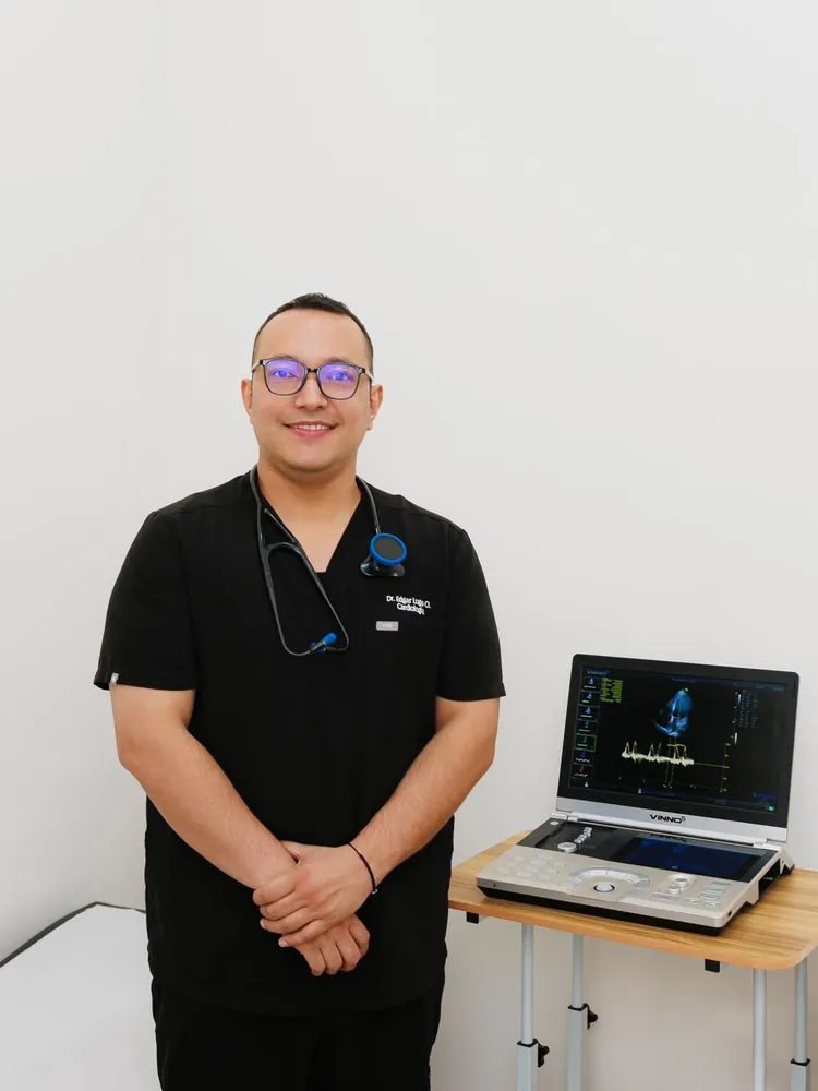 Dr. Edgar Lugo Osuna con uniforme negro, de pie junto al ecocardiógrafo de su consultorio en Nogales